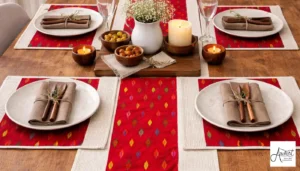A rustic wooden table set with red ikat patterned handmade placemats, white ceramic plates, linen napkins with wooden cutlery, and white floral centerpieces with candles.