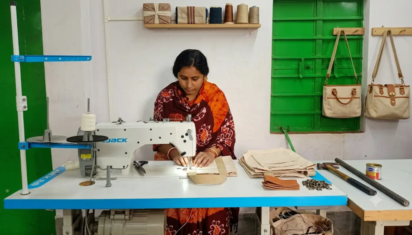 A woman carefully stitching fabric panels at a workshop table with finished tote bags hanging on the wall during a handmade bag journey.